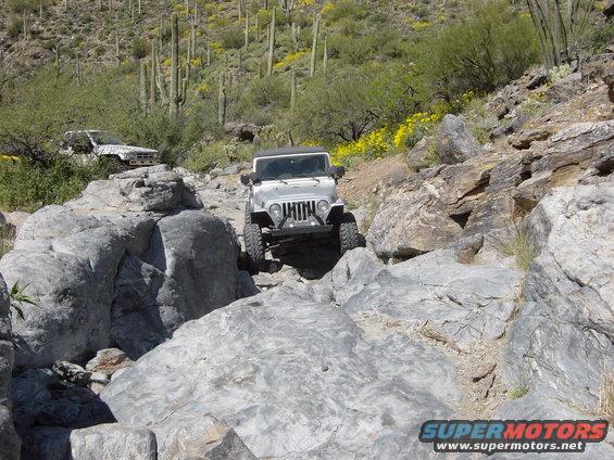 Silver Jeep Wrangler navigating a rocky trail with a white SUV in the background