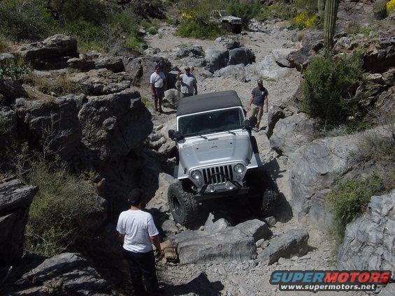 Silver 2004 Jeep Wrangler navigating a rocky off-road trail with people watching. Another Jeep is visible further up the trail