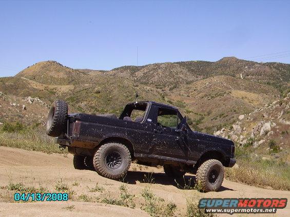 Black 1990 Ford Bronco with spare tire on back, driving on dirt trail in hilly terrain
