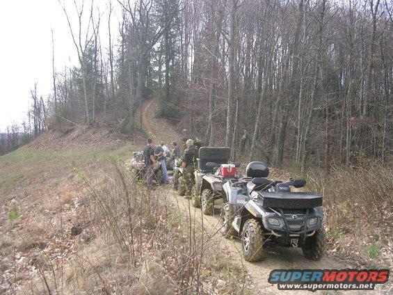 Several ATVs parked on a dirt trail with people standing around them, surrounded by trees