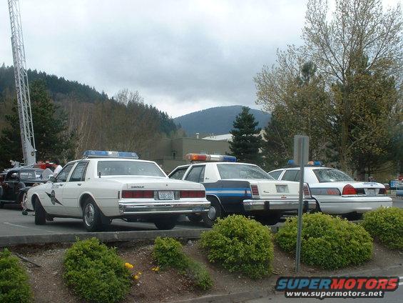 Three white police cars parked in a lot, one with a ladder truck behind it