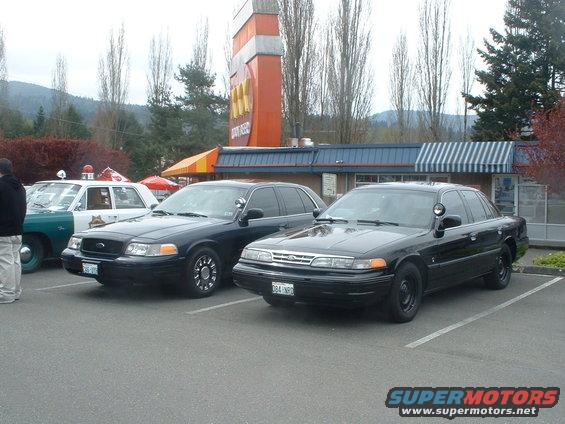 Two black police sedans parked side-by-side in a parking lot; one white and green police car is partially visible to the left