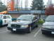 1987 Ford Crown Victoria LTD - A dark police car is parked in a lot next to a classic police car, with a building and trees behind them.