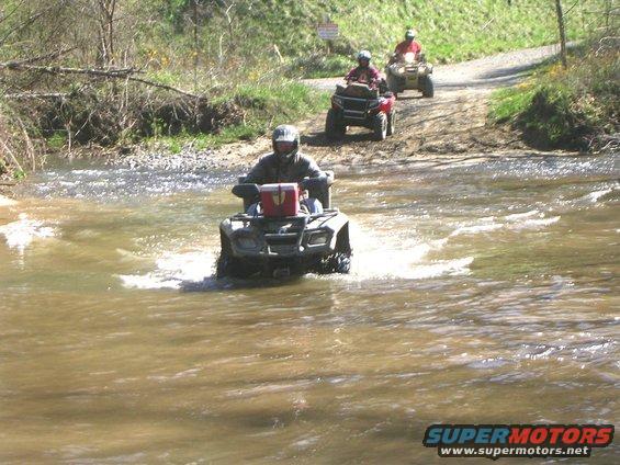 Front view of a four-wheeler crossing a stream, with two others behind