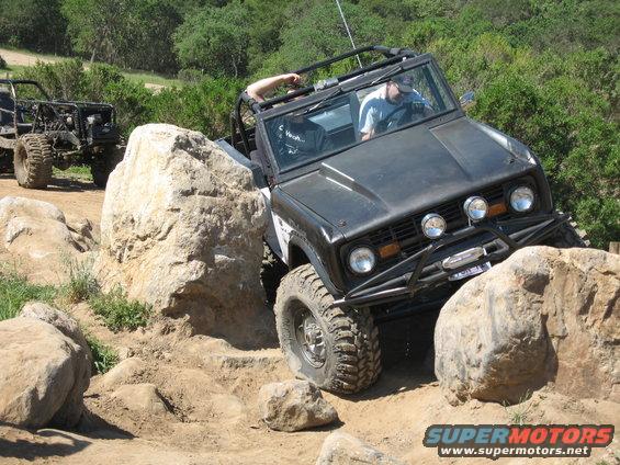 Black off-road vehicle with roll cage climbing over rocks, another off-road vehicle behind it