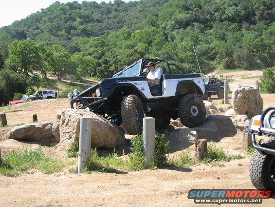 Off-road vehicle with large tires climbing over rocks on a dirt trail. Another vehicle is visible in the background