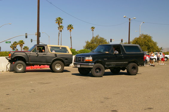 Two Ford Broncos parked on a street, one dark gray with tan top, one black. Palm trees and traffic lights in background