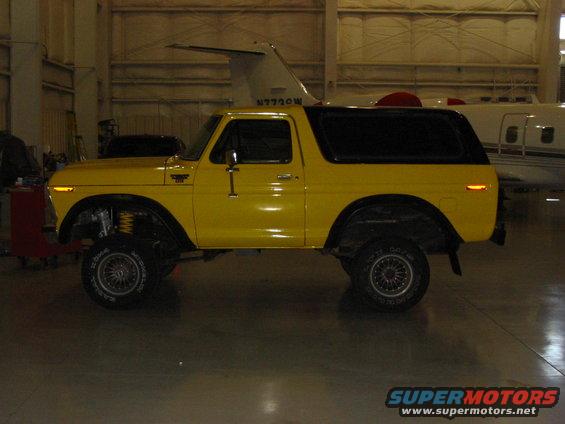 Yellow 1978 Ford Bronco side view, lifted suspension, black hardtop, parked indoors near aircraft