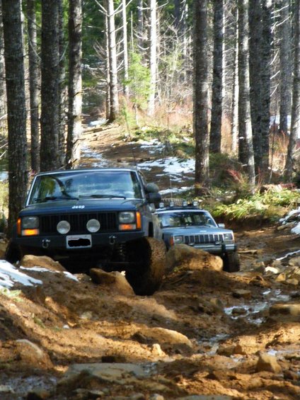 Two Jeep Cherokees navigating a muddy, wooded trail with snow patches