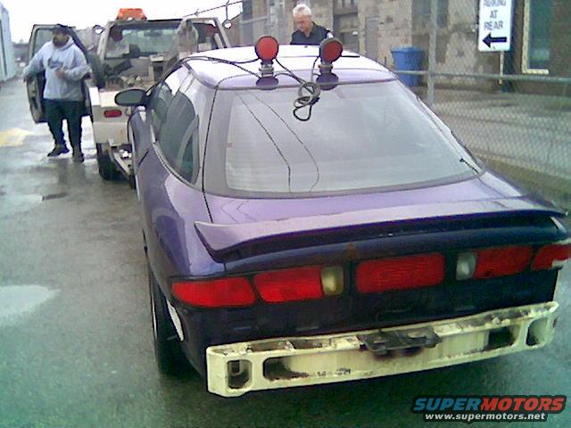 1996 Ford Probe being towed by a flatbed truck, rear view. Tow truck driver stands by the car door