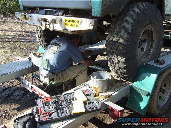 Guy working on the rear axle of a lifted off-road vehicle on a trailer. Tools and a beer are nearby