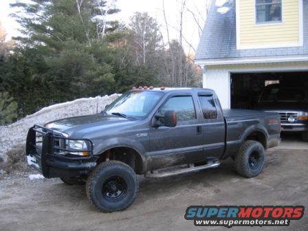 Dark gray Ford F-250 truck with a brush guard parked outside a garage. Another truck is visible inside the garage