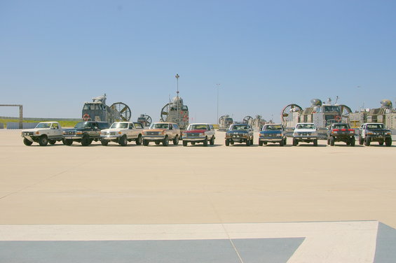 Line of 1986 Ford Bronco SUVs and Ford F-Series trucks parked on pavement with hovercraft in background