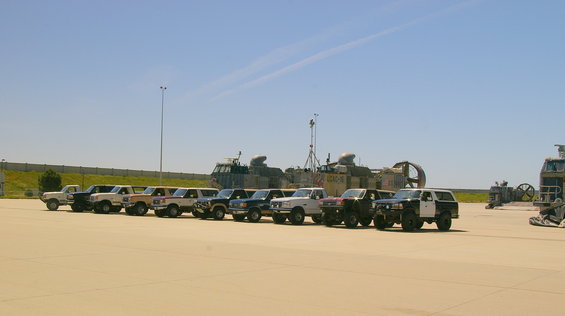 Multiple SUVs and trucks parked in a line on an airfield with hovercraft in the background