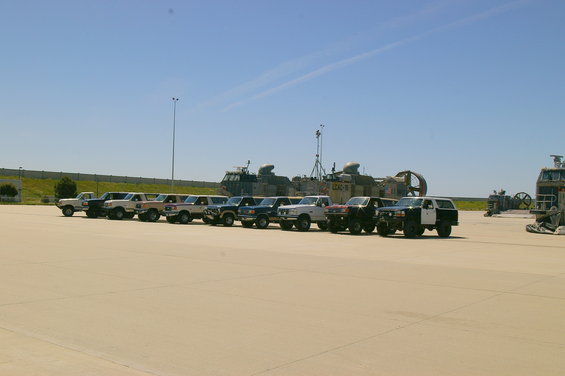 Several SUVs and trucks parked in a line on pavement with large boats in the background