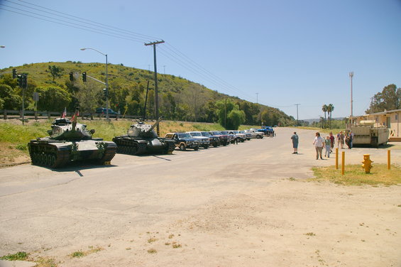 Two tanks parked on a road with a line of SUVs behind them