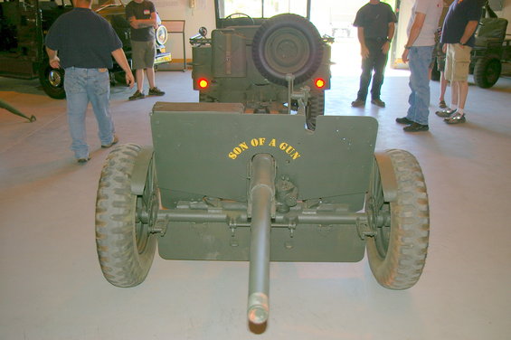 Rear view of a green military artillery piece with 'SON OF A GUN' painted on it, parked indoors near other vehicles and people