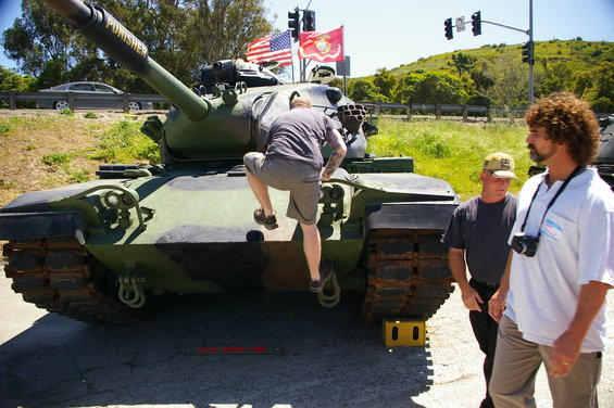Camouflaged tank with 'PUNISHER' on barrel, a man climbing on front, and two men standing nearby
