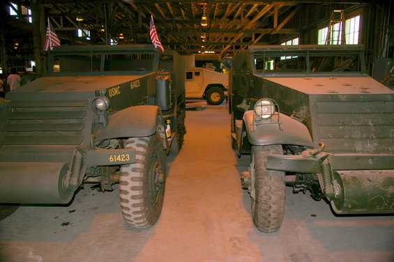 Two olive drab military half-track vehicles parked side-by-side in a hangar, with a white Humvee visible behind them