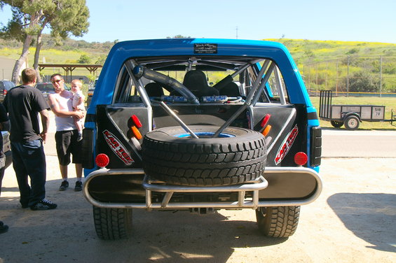 Blue off-road SUV with roll cage and spare tire mounted on the back. People stand nearby