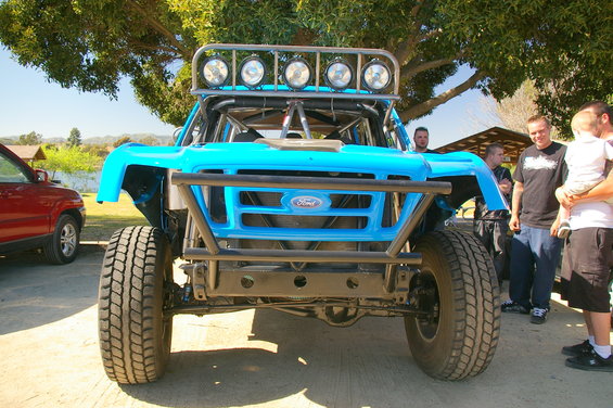 Front view of a blue off-road truck with a roll cage and lights on top. A red SUV is parked to the left. People stand to the right