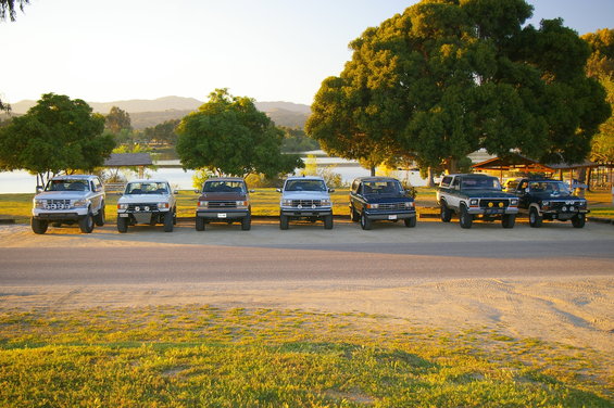 Seven SUVs parked in a line facing front, with a lake and trees in the background. The first vehicle appears to be a 1986 Ford Bronco