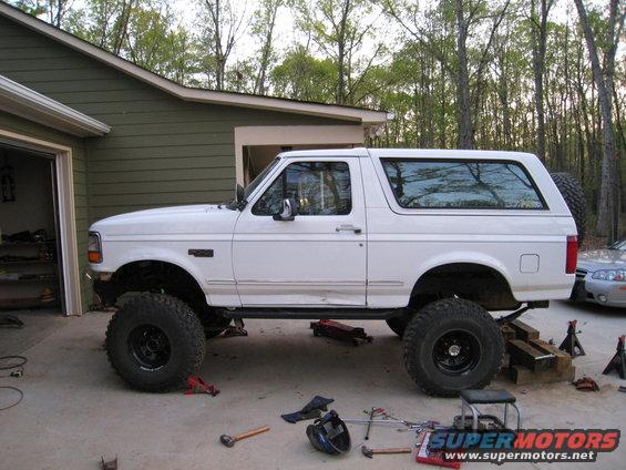 White Ford Bronco on jack stands with large tires, parked in a driveway next to a garage