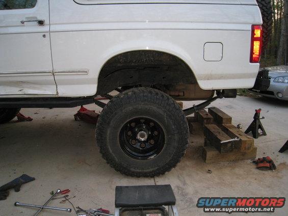 White Ford Bronco on jacks with large tires and suspension work. Another car is visible in the background