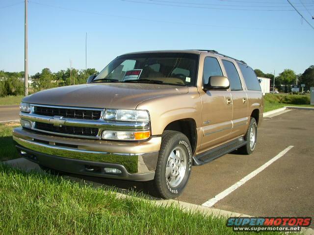 Tan Chevrolet Suburban parked in a lot. Has a 'For Sale' sign in the window