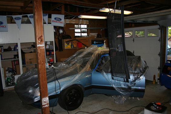 1990 Ford Mustang covered in plastic wrap in a garage, with a grille hanging nearby