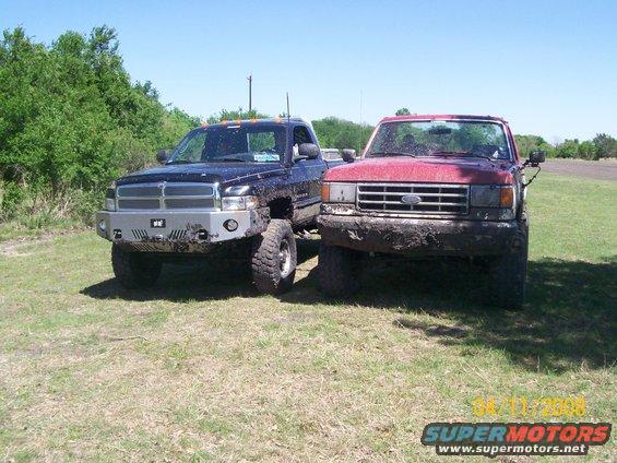 Muddy red Ford truck next to a muddy black Dodge truck, both on grassy field