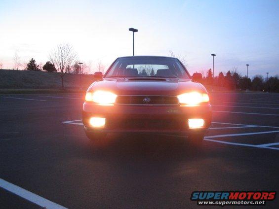 Front view of a red 1997 Subaru Legacy with headlights on in a parking lot at dusk