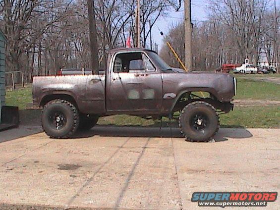 Rusty pickup truck with large tires, parked outside. Another white truck is visible in the background