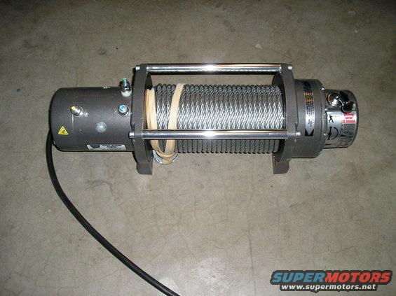 Gray electric winch with steel cable wound on drum, metal frame, and black power cord