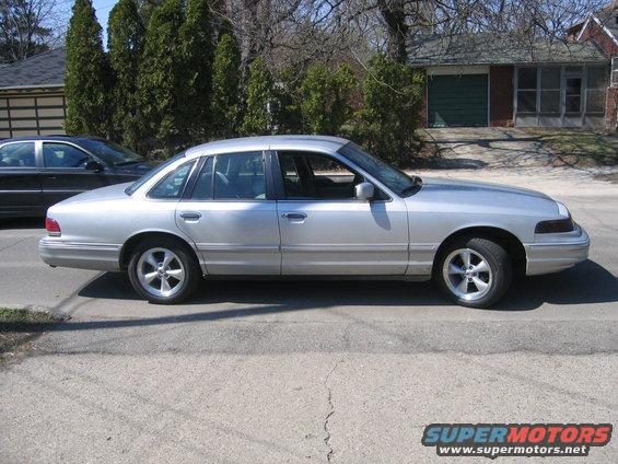 Side view of a silver 1996 Ford Crown Victoria parked on a street. Another car is behind it