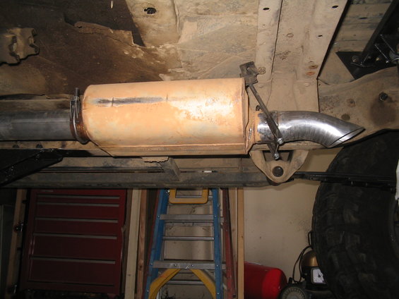 Underside view of a vehicle's exhaust system with a rusty muffler and chrome tailpipe. A red toolbox and ladder are visible in the background