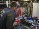 1979 Ford Bronco - Two men work on a metal component in a cluttered workshop setting, with tools and equipment visible in the background.