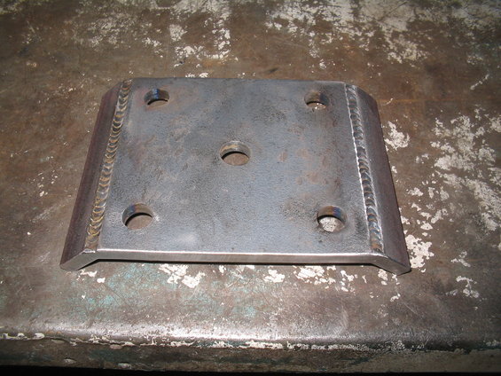 Metal plate with four outer bolt holes and one central hole, showing visible weld marks along the edges