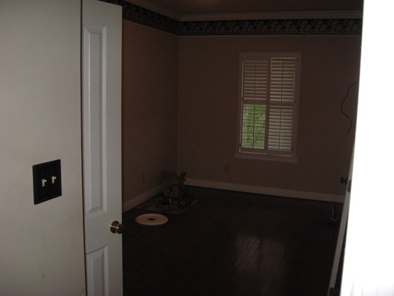 Interior of a room with a closed door on the left, a window with blinds, and dark wood floors. Some items are on a rug in the corner