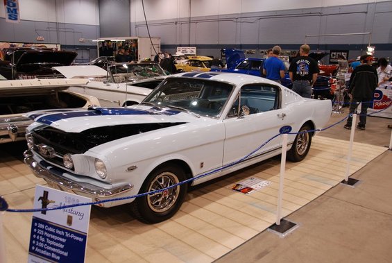 White 1966 Ford Mustang with blue stripes, hood open, on display. Other cars visible in background
