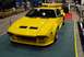 1996 Ford Bronco - A bright yellow sports car with a prominent rear wing is displayed indoors, with other vehicles and exhibits visible in the background.