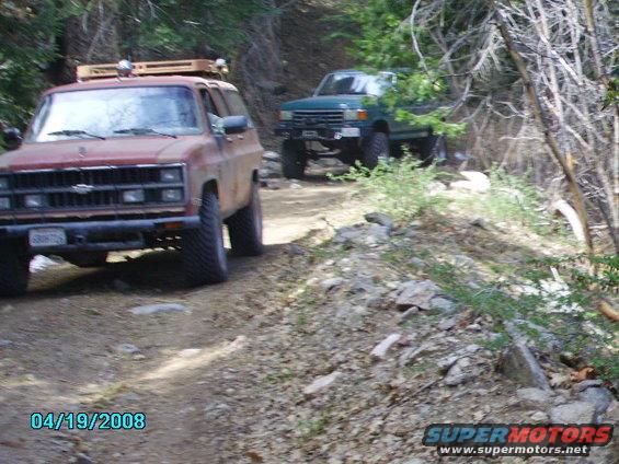Red SUV with roof rack on dirt road, green truck behind