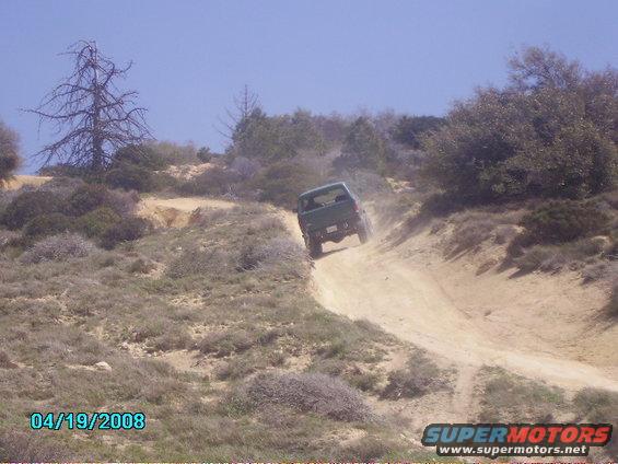 Green SUV driving up a dusty dirt road on a hill. There's a dead tree on the left