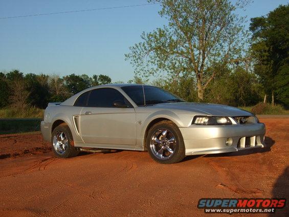 Silver Ford Mustang parked on dirt, front 3/4 view, chrome wheels, clear sky