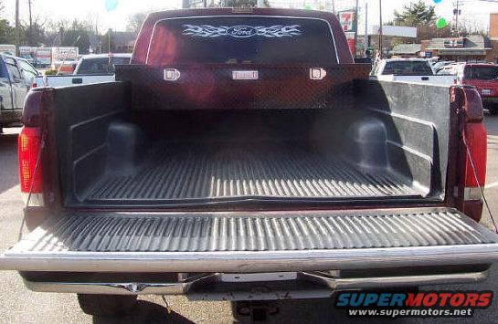Red pickup truck bed with a toolbox mounted above it. Other vehicles and buildings in background