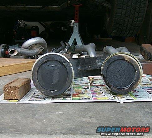 Two engine pistons on newspaper, with exhaust pipes and turbocharger parts in background
