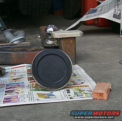 Round automotive part with black surface, resting on newspaper. Metal hitch ball and bracket visible behind it