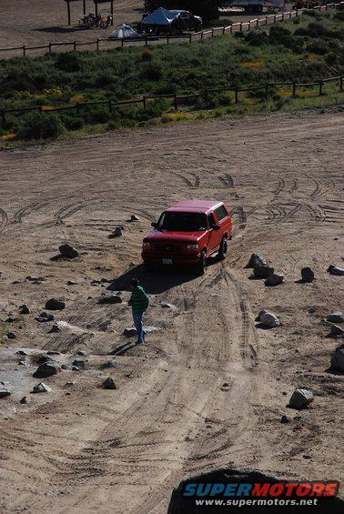 Red 1995 Ford Bronco driving on a dirt road with tire tracks, a person stands nearby