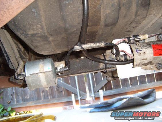 Underside of a vehicle showing the fuel filter and part of the fuel pump assembly. There's a large fuel tank above