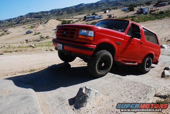 Red Ford Bronco with black wheels on a rocky incline, front-left tire lifted